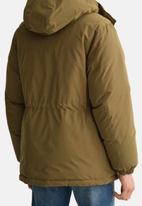 Olivengrøn dunjakke med hætte, der har en let indsnævret talje, elastiske manchetter og en glat, mat tekstur. Forstærkede syninger er synlige.