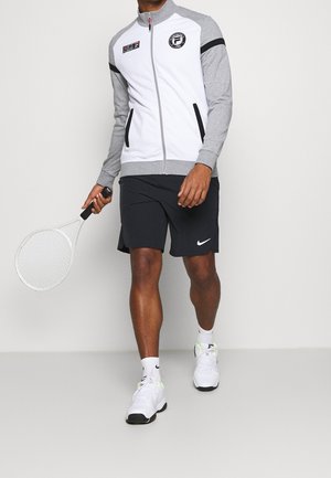 Homme portant une veste de sport zippée grise et blanche, un short noir, des baskets blanches Nike, tenant une raquette de tennis blanche, marchant sur un fond uni.
