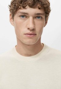 Cream-colored, textured knit sweater with a round neck. Close-up of a male model with curly hair and blue eyes against a plain background.