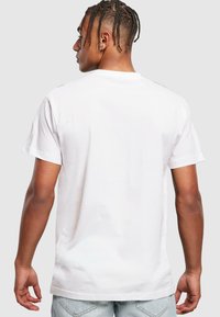 T-shirt en coton blanc à manches courtes, col rond et texture lisse. Modèle vu de dos, avec des cheveux tressés et une coupe décontractée.