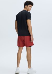 Camiseta negra de manga corta combinada con pantalones cortos deportivos rojos. El modelo lleva zapatillas blancas, mostrando un aspecto casual y deportivo por detrás.