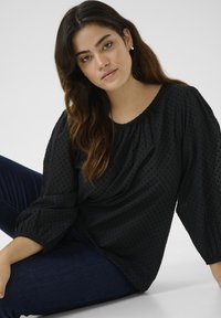 Zwarte blouse met een gestructureerd patroon, ronde halslijn en volumineuze mouwen. Gecombineerd met donkerblauwe jeans zit het model tegen een neutrale achtergrond.