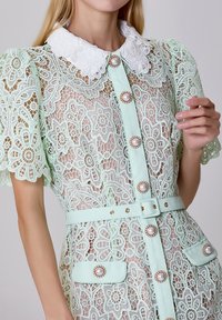 Robe en dentelle vert menthe avec un col en dentelle blanche, des boutons décoratifs en perles et une ceinture assortie. Présente des manches courtes bouffantes et des poches.