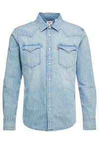 Ljusblå denimskjorta med spetsig krage, två bröstfickor och silverfärgade tryckknappar. Har en något urblekt textur och rundad nederkant.