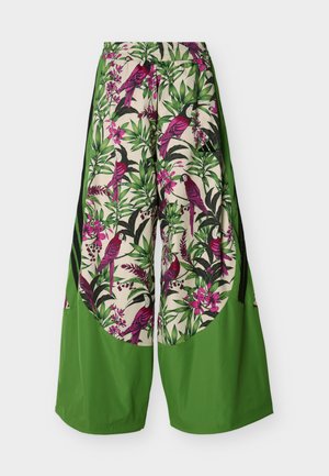 Pantalones de pierna ancha con paneles inferiores sólidos en verde y secciones superiores que presentan loros rosas y hojas tropicales verdes sobre un fondo beige.