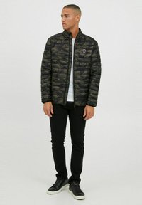 Veste rembourrée camouflage matelassée avec un col montant, fermeture éclair avant et détail de logo sur la poitrine, associée à un pantalon noir et des baskets.