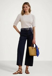 Žena v bielom tričku s krátkym rukávom, čiernych širokých nohaviciach s opaskom, hnedých sandáloch a s ratanovou kabelkou, do ktorej je zaviazaná modrá šatka.