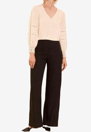 Nainen, joka pukeutunut kermaiseen pitkähihnaiseen paitaan, jossa on pitsiyksityiskohtia, ja mustiin leveälahkeisiin housuihin, yhdistettynä mustiin terävikärkiisiin tasapohjaisiin kenkiin.