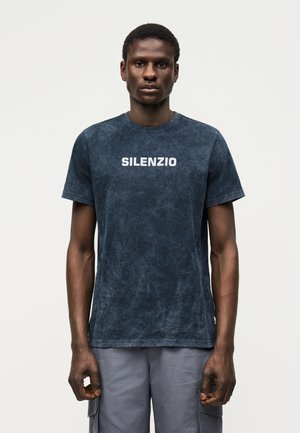 Άνδρας που φοράει μια σκούρα μπλε ξεθωριασμένη μπλούζα με την επιγραφή «SILENZIO» στο στήθος και γκρι παντελόνι φόρμας, όρθιος μπροστά σε απλό φόντο.