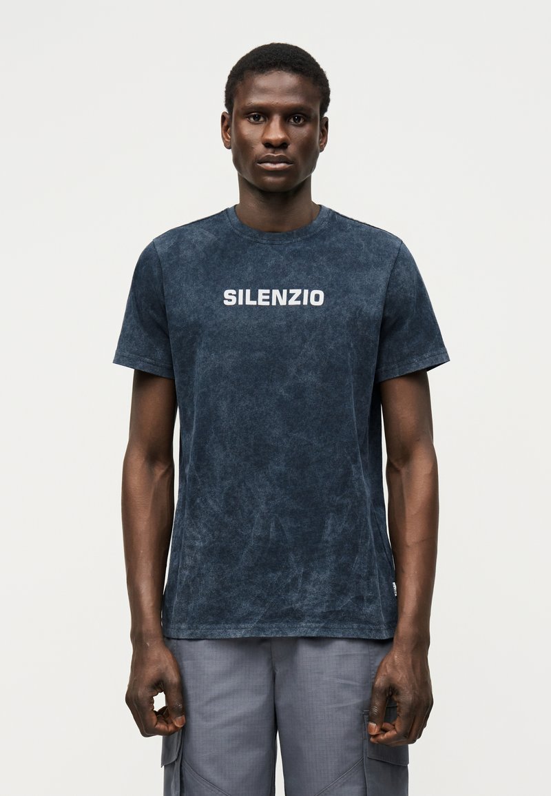 Άνδρας που φοράει μια σκούρα μπλε ξεθωριασμένη μπλούζα με την επιγραφή «SILENZIO» στο στήθος και γκρι παντελόνι φόρμας, όρθιος μπροστά σε απλό φόντο.