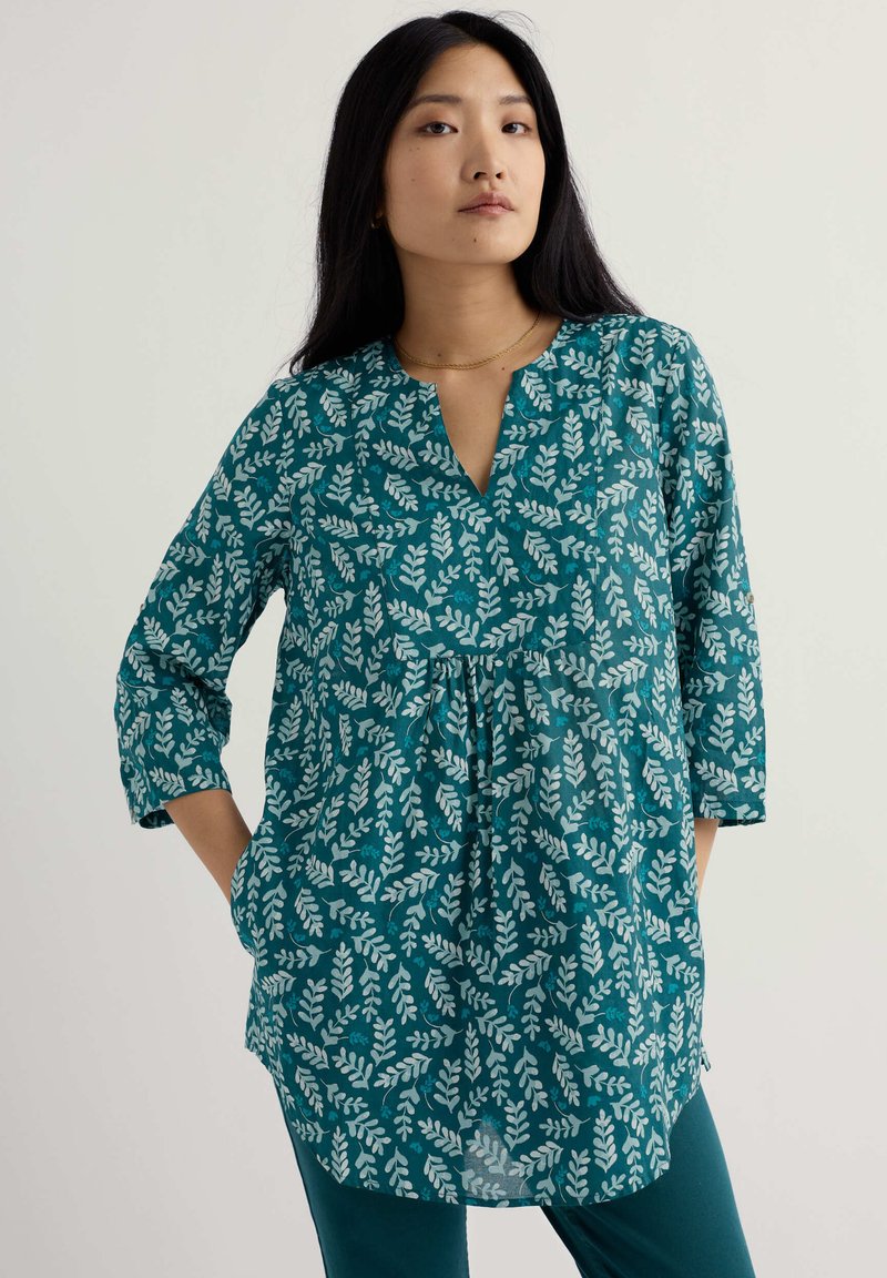 Frau mit langen dunklen Haaren, die eine türkisfarbene Tunika mit hellem Blattmuster und türkise Hosen trägt, steht mit den Händen in den Taschen vor einem schlichten Hintergrund.