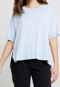 Světle modrý krátký svetr s uvolněným střihem, žebrovaným lemem a postranními rozparky. Vyroben z měkké tkaniny s hladkou texturou.