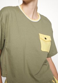 Osoba v olivově zeleném tričku s krátkým rukávem a límcem s žlutým okrajem, se žlutou kapsou na knoflík na hrudi, stojící na bílém pozadí.