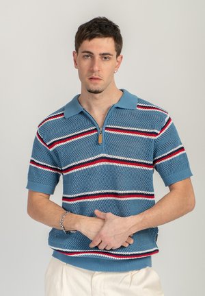 Jeune homme aux cheveux courts portant un polo en maille bleu avec des rayures rouges, blanches et noires, debout les mains jointes devant un fond uni.