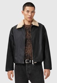 Chaqueta bomber negra con un cuello de borreguillo beige, cierre frontal con cremallera y mangas largas, combinada sobre un suéter marrón texturizado, llevada con jeans oscuros.