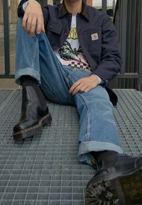 Schwarze Leder Chelsea-Stiefel mit strukturiertem Außensohlen, blaue Jeans, die an den Säumen hochgerollt sind, und eine marineblaue Jacke über einem Grafik-T-Shirt.