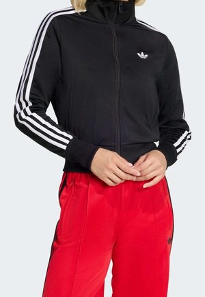 Osoba nosząca czarną kurtkę zapinaną na zamek marki Adidas z białymi paskami na rękawach oraz czerwone spodnie dresowe Adidas z czarnymi paskami po bokach.