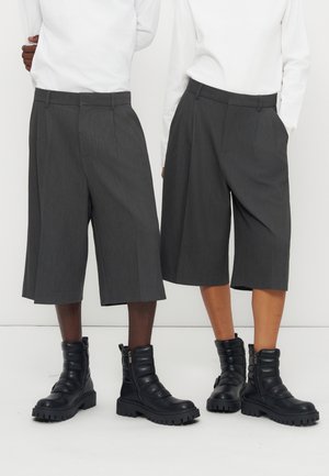 Twee personen dragen witte tops, donkergrijze wijde knielange broeken en zwarte dikke schoenen, staand naast elkaar tegen een witte achtergrond.