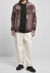 Man som bär en brunmönstrad zip-up-jacka, svart skjorta, krämfärgade cargobyxor och svarta sneakers mot en plain grå bakgrund.