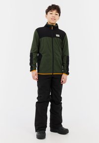 Fleecejack met een donkergrijze en zwarte kleur, voorzien van een rits aan de voorkant, hoge kraag en oranje accenten. Gecombineerd met zwarte waterdichte broek en laarzen.