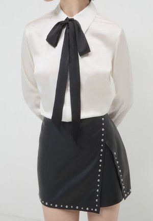 Persona che indossa una blusa di raso bianca con un grande fiocco nero e una minigonna di pelle nera ornata da borchie argentate lungo i bordi.