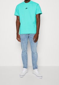 Turkoosfärgad t-shirt i bomull med rund halsringning och liten svart Nike-logotyp, kombinerad med ljusblå slitna jeans och vita sneakers.