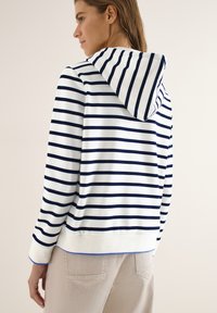 Femme portant un sweat à capuche blanc avec des rayures horizontales bleu marine, vue de dos, associée à un pantalon beige clair.