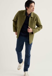 Olívazöld kabát elülső zsebekkel, tengerészkék póló fölött viselve, kék nadrággal és bézs részletekkel díszített fehér tornacipővel párosítva.