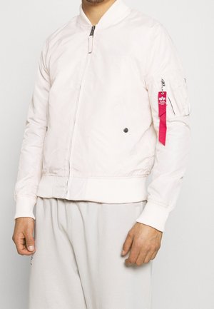 Mężczyzna ubrany w białą kurtkę bomber z zamkiem z przodu i czerwoną naszywką na kieszeni na lewym rękawie na jednolitym tle.