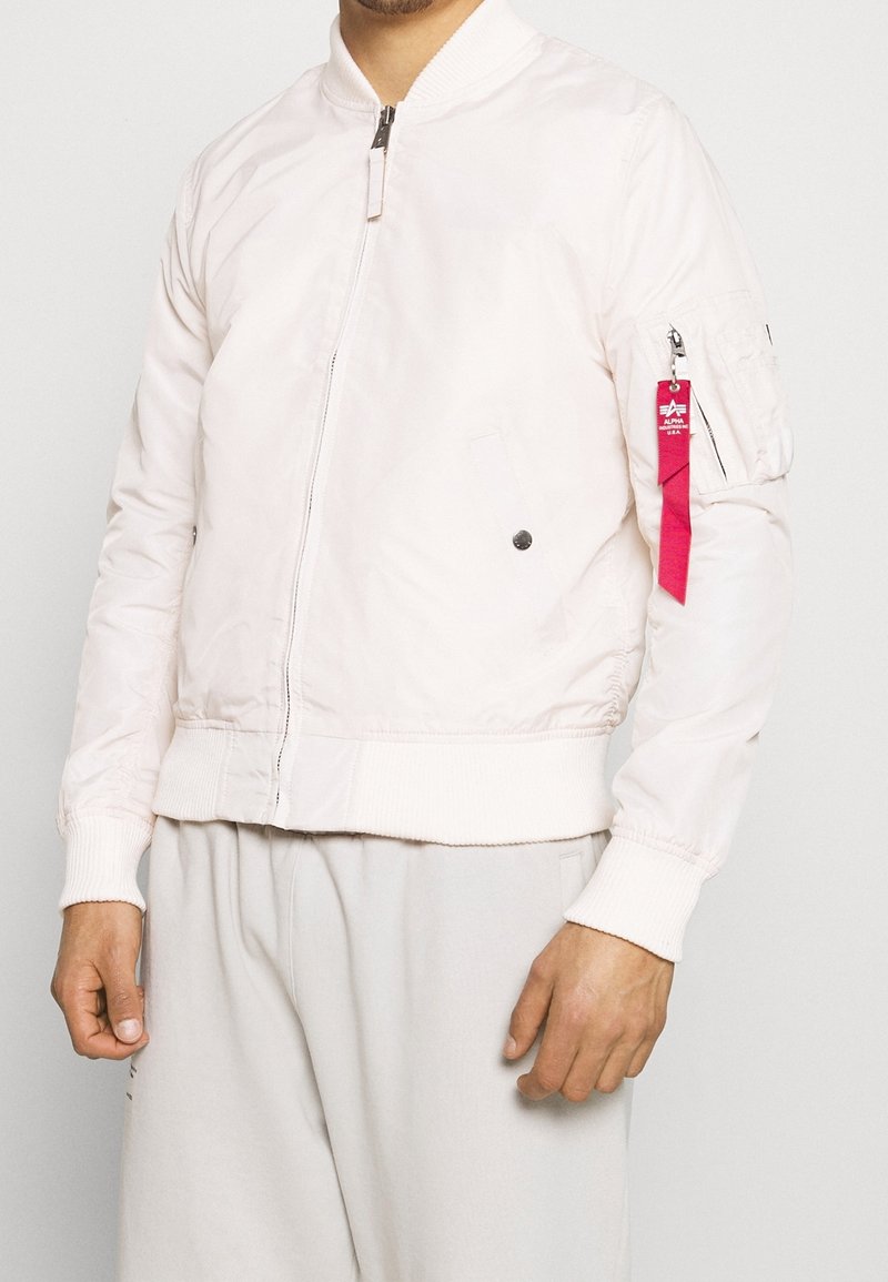 Homme portant une veste bomber blanche avec une fermeture éclair à l'avant et une étiquette rouge sur la poche de la manche gauche, devant un fond uni.