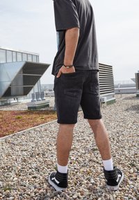 Shorts de mezclilla negros con dobladillos enrollados, una camiseta gráfica y zapatillas altas negras con acentos blancos, ubicados en una azotea de gravilla.