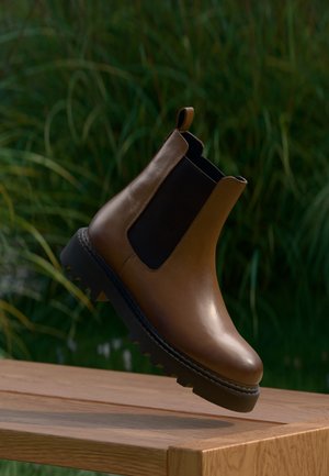 Botte Chelsea en cuir marron avec panneaux latéraux élastiques noirs et semelle noire épaisse en lévitation au-dessus d'une surface en bois avec un fond de verdure.