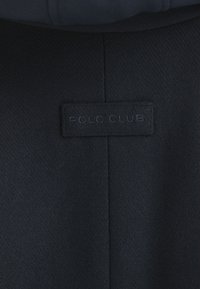 Navyblått tyg i nära bild med en strukturerad yta, med en sydd "POLO CLUB"-etikett i en matchande mörk ton.