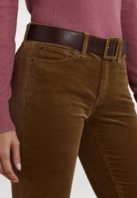 Hnědé manšestrové kalhoty s žebrovanou texturou, doplněné tmavě hnědým koženým pásem s dekorativní sponou. Viditelný růžový top s dlouhým rukávem.