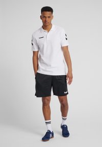 Witte poloshirt met zwart logo en zwarte chevrons, gecombineerd met zwarte shorts met witte branding. Model draagt blauwe sportschoenen en gestreepte sokken.