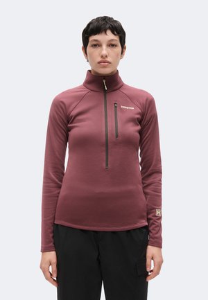 Persona joven con cabello corto que lleva una chaqueta con cremallera Patagonia de color granate y pantalones negros sobre un fondo claro y sencillo.