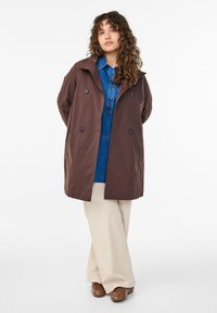 Brauner Mantel mit doppelter Knopfleiste und großem Kragen, mit Knopfdetails, kombiniert mit einem Jeanshemd und cremfarbenen, weit geschnittenen Hosen. Braune Schuhe vervollständigen den Look.