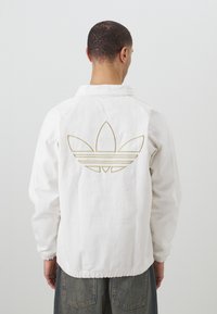 Bílá bunda s límečkem, na zadní straně se zlatým logem Adidas. Elastické manžety a spodní lem, vyrobena z odolné látky.