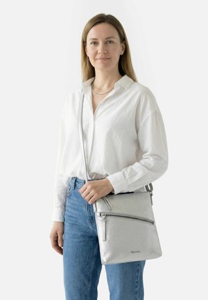 Frau in weißem Hemd und blauer Jeans, die eine silberne Umhängetasche mit vorderer Reißverschlusstasche trägt, vor einem schlichten hellen Hintergrund stehend.