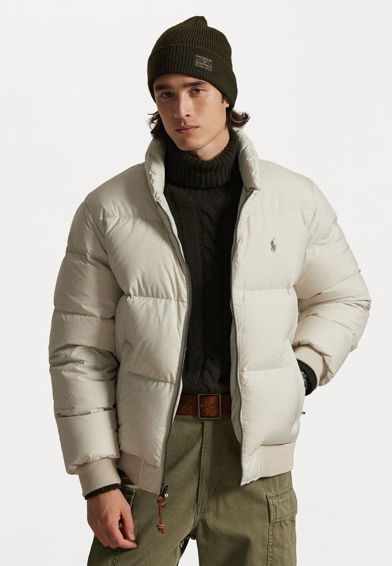 Casaco puffer, bege claro, design acolchoado. Punhos canelados, fecho de zip. Usado sobre um suéter de malha em verde-escuro, calças cargo em oliva e um gorro escuro.