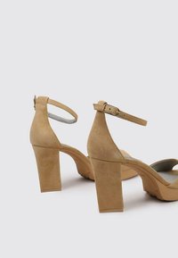 Sandalias de tacón de ante beige con un amplio tacón de bloque, correa al tobillo y diseño de punta abierta. Presenta una superficie texturizada y una suela resistente.