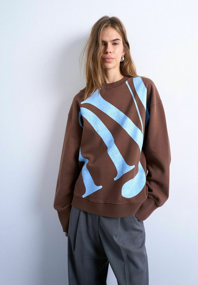 Jeune femme aux longs cheveux portant un sweat-shirt marron avec un grand motif abstrait bleu clair et un pantalon plissé gris, debout contre un mur blanc.