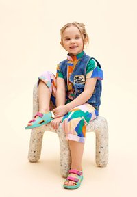 Chaleco de mezclilla sobre una camisa de colores con cuello, combinado con pantalones cortos con estampados y sandalias color teal con tiras rosas, sentado en un taburete texturizado.