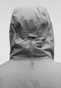 Tagantvaade isikule, kes kannab heledat halli kapuutsiga jakki, mille kapuutsi pingutamiseks on reguleeritav nöör.