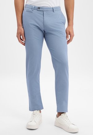 Uomo che indossa pantaloni aderenti azzurri chiaro e sneakers basse bianche, in piedi contro uno sfondo bianco uniforme.