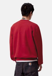 Felpa rossa con colletto a girocollo e maniche lunghe, caratterizzata da una superficie liscia. Indossata sopra pantaloni scuri a fantasia, con un'etichetta del logo visibile sulla tasca.