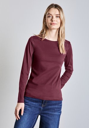 Top lungo bordeaux a maniche lunghe con un design aderente e scollatura rotonda. Abbinato a jeans in denim blu. Morbido al tatto, con dettagli minimi.