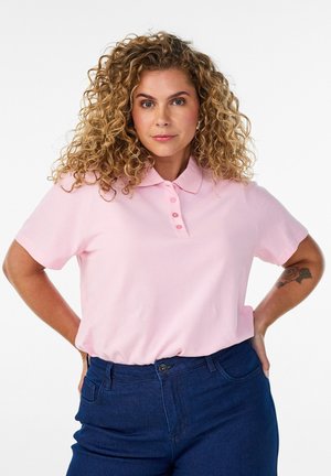 Žena s kučeravými blond vlasmi nosí ružové polotričko a tmavomodré nohavice, pózuje s rukami v bokoch na bielom pozadí.