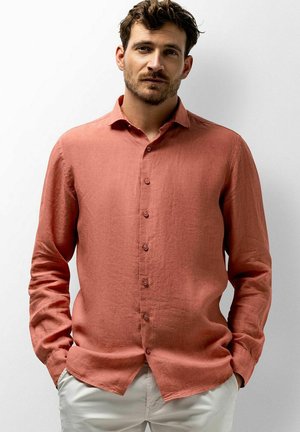 Jonge man met kort bruin haar, gekleed in een roestkleurig overhemd met lange mouwen en lichtbeige broek, handen in de zakken, neutrale achtergrond.