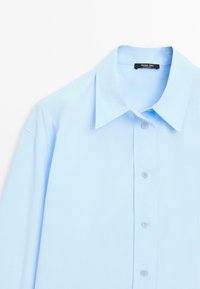 Camicia azzurra chiara con colletto a punta, maniche lunghe e etichetta del marchio visibile all'interno del colletto.
